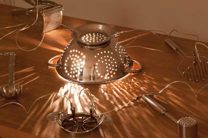 Installation aus Küchenutensilien auf einer Holzplatte, bestehend aus einem Nudelsieb, einer Käsereibe, einem Nudelheber, einem Kartoffelstampfer und einem Fleischklopfer, um nur die Utensilien auf dem Foto zu nennen. Alle sind mit Elektrokabeln verbunden, sodass ein Stromkreislauf entsteht und somit die Glühbirne im Nudelsieb zum Leuchten gebracht werden kann. Mona Hatoum, Sammlung Goetz München