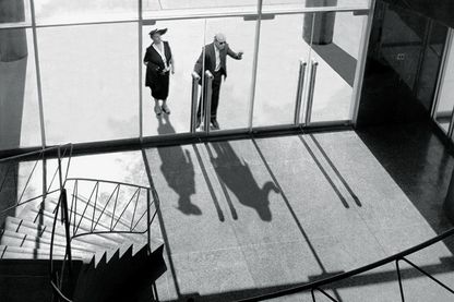 Video Still in schwarzweiß, auf dem ein Mann und eine Frau zu sehen sind, die von einer Wendeltreppe im Inneren eines Gebäudes aus gesehen vor dem verglasten Eingang und in den Raum schauen. David Claerbout, Sammlung Goetz München