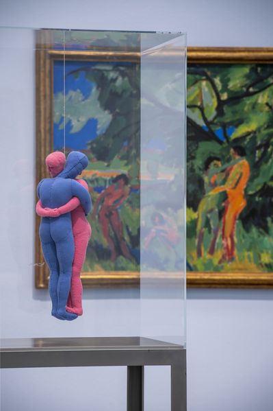 Skulptur einer rosafarbenen und einer hellblauen Stoffpuppe, die sich umarmen in einem Plexiglaskasten. Dahinter hängt eine expressionistischer Malerei in kontrastreichen Farben (grün, blau, rot, orange) mit vier nackten Menschen vor einer Landschaft. Darunter ein Mann und eine Frau, die sich umarmen
