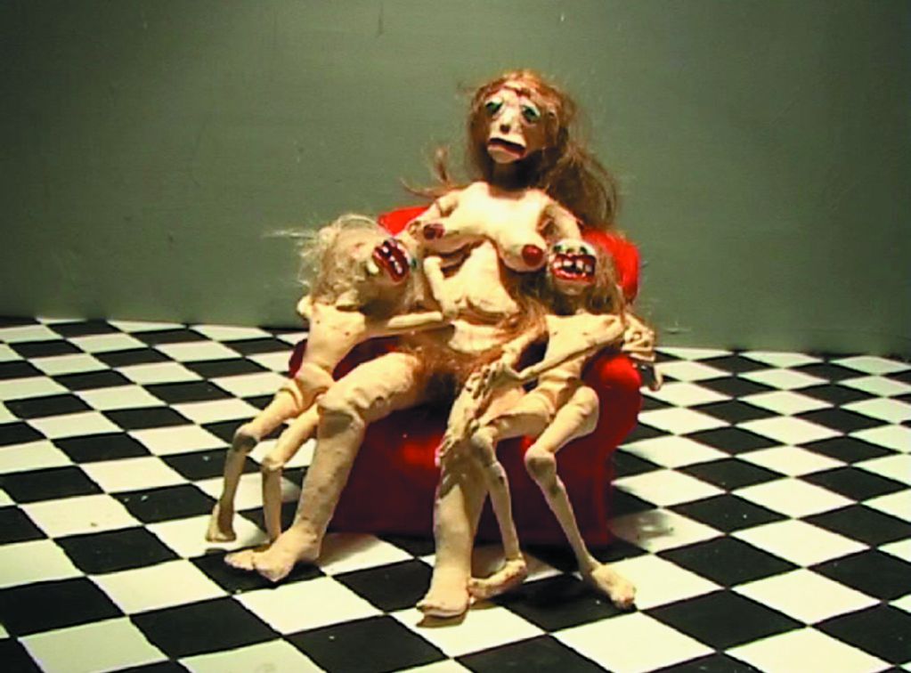 Video Still that shows three figures made of plasticine. They are a naked mother figure sitting on a red armchair, flanked by her two daughters, who have grotesquely large mouths. The floor is tiled in black and white. Nathalie Djurberg, Sammlung Goetz Munich