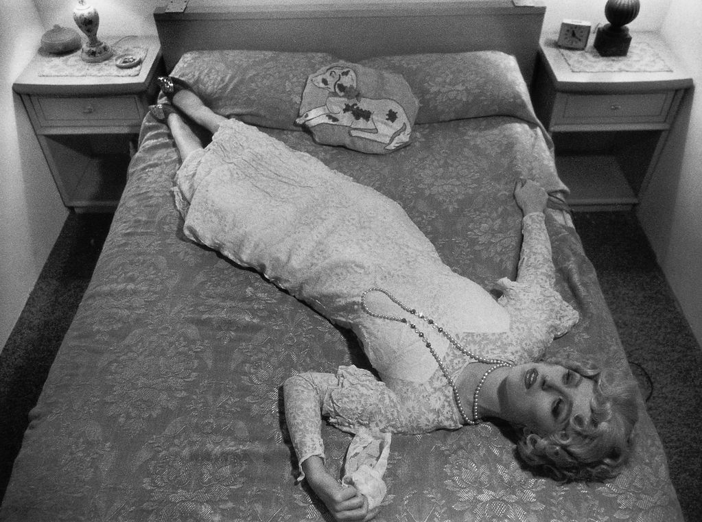 Black and white photography, a woman with a white lace dress and pearl necklace lies diagonally on a bed, her shoes on a pillow. In her left hand she is holding a handkerchief, with her right hand she is clutching the bedcover.  Cindy Sherman, Sammlung Goetz, Munich