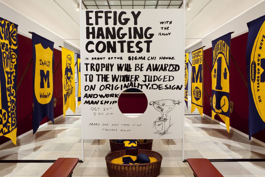 Installation consisting of 13 felt banners with poles, two framed pictures, a wooden board painted on two sides, two dog baskets, a basketball, two footballs and a felt cushion. The installation is meant to give the appearance of an alma mater (American university) hall of fame and is painted in the colours purple and yellow. Mike Kelley, Sammlung Goetz Munich