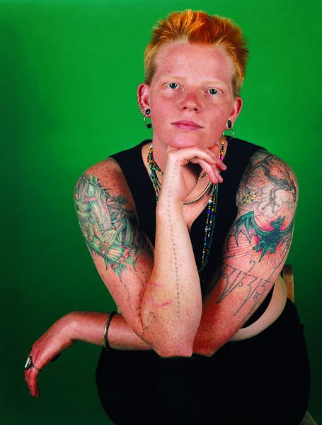 Here is a portrait photograph of a red-haired woman with short haircut and tattooed upper arms in front of a green background. She is sitting on a chair, her legs crossed, her elbows leaning on her knees, one hand on her chin and looking directly into the camera.