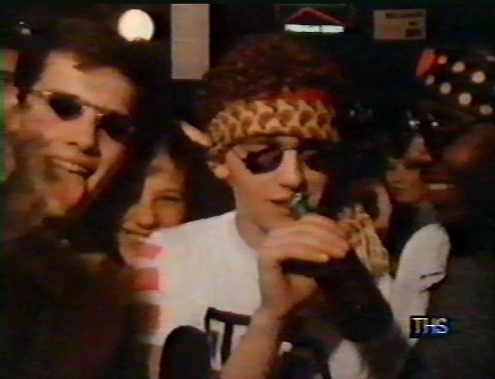 Group of young people partying. They have sunglasses on and bandanas tied around their heads. On the left, a young man sticks his tongue out at the camera/viewer, on the right another sips at a bottle