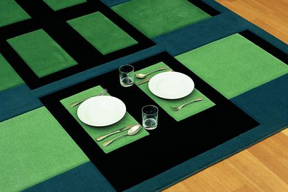 Diese Ausstellungsanischt zeigt einen Teppich der Künstlerin Andrea Zittel, auf dem man (zu Abend) essen kann. Er besteht aus moosgrünen, blauen und schwarzen, rechteckigen Formen, in denen zwei Teller, Bestecke und Gläser gegenüber voneinander aufgestellt sind.