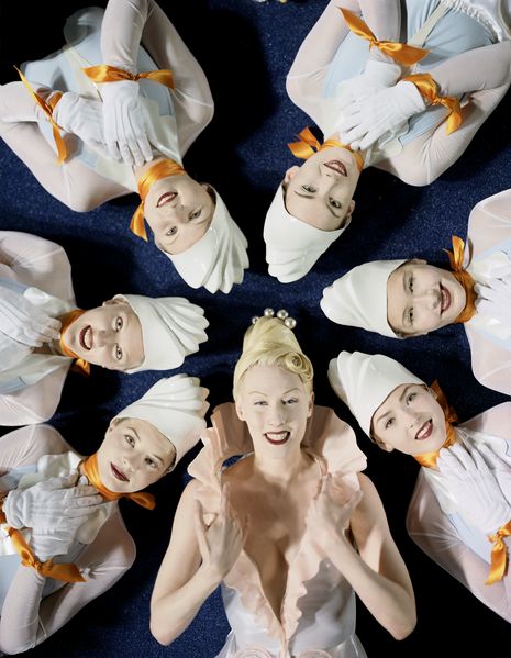 This photograph shows seven white women lying on the ground in a circle. The detail of the photograph shows them from the head to the chest. All the women are wearing very bright makeup and white clothes. The woman at the bottom center has blonde white hair, while the other women are wearing a white plastic hood that looks like a icing on the cake. Matthew Barney, Sammlung Goetz Munich 