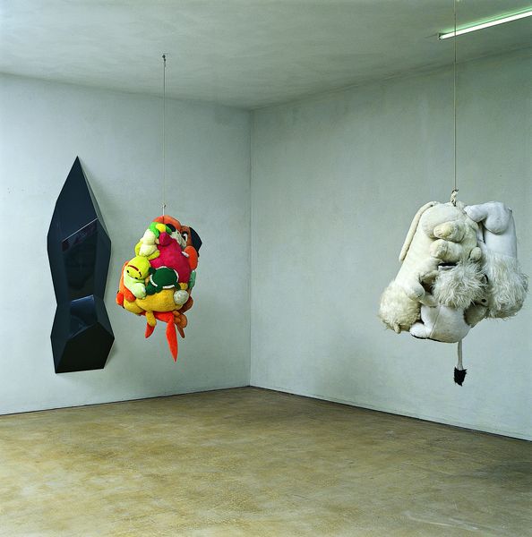 This installation view shows a black, smooth and shiny wall object and intertwined stuffed animals dangling from the ceiling.
