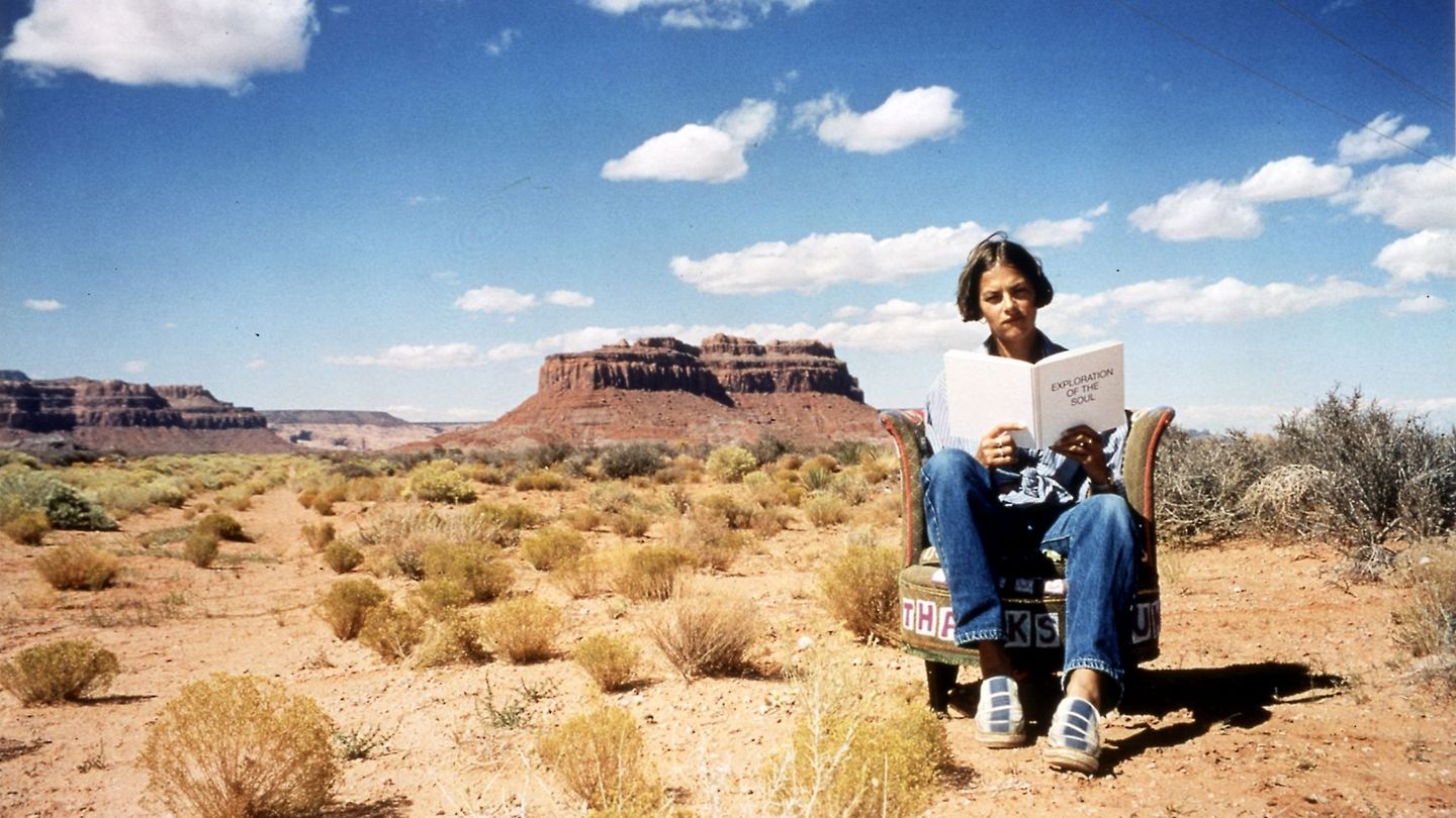 Photography of the artist in Monument Valley on an armchair embroidered by her with a self-written publication in her hand.