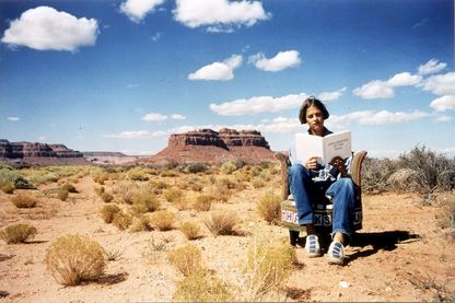 Photography of the artist in Monument Valley on an armchair embroidered by her with a self-written publication in her hand.