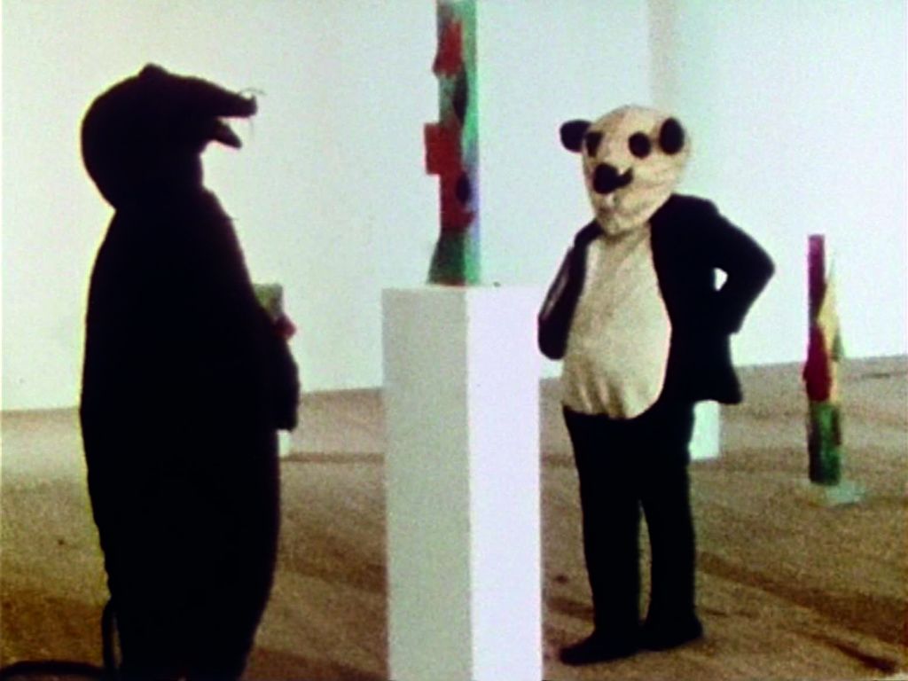 Video Still showing two people dressed up like mice in an exhibition room, attentively observing a contemporary coloured sculpture on a white plinth. Peter Fischli/David Weiss, Sammlung Goetz Munich