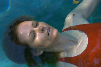 Video Still einer Frau, die in dem türkisblauen Wasser eines Swimming Pools zu schweben scheint. Sie hat weiße Haut, brünettes Haar und feine Gesichtszüge und trägt einen tomatenroten Badeanzug. Teresa Hubbard/Alexander Birchler, Sammlung Goetz München