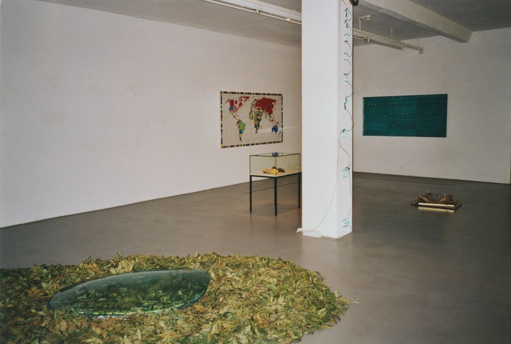 Here you can see a view of the exhibition with a floor sculpture of laurel leaves and a glass pane in the foreground. An embroidered wall object with a world map by Alighiero Boetti is on the left white wall, in a showcase in front of it is a pair of gold-plated shoes by Jannis Kounellis. At the top of the column in the middle of the room there is a preparated caiman with neon numbers below, while on the back wall there is a three-part wall object with ballpoint pen strokes and letters. Underneath is a sculpture made of gilded picture frames, cushions and stone.