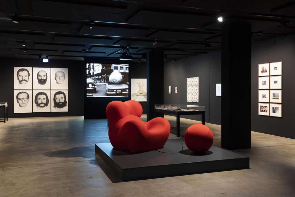 Ausstellungsansicht mit einem roten, knubbeligen Sessel samt Fußablage im Fokus. Dahinter sind einige Werke an den schwarzen Wänden sowie eine Videoprojektion zu erkennen. Gaetano Pesce, Sammlung Goetz München