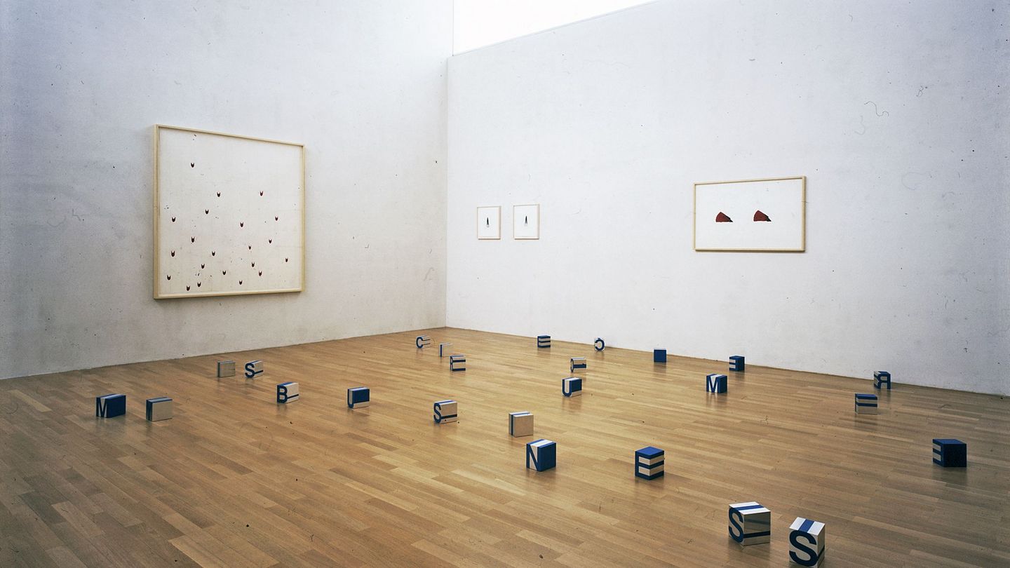 This installation photograph shows a total of four works by the artist Roni Horn. The approximate centre of the picture forms a corner of the room, on one wall is a large-format painting, on the other wall are four smaller works on paper. Small cubes with letters are spread out on the wooden floor.