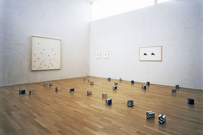 Diese Installationsaufnahme zeigt insgesamt vier Arbeiten der Künstlerin Roni Horn. Die ungefähre Bildmitte bildet eine Raumecke, an der einen Wand ist eine großformatige Malerei, an der anderen Wand vier kleinere Papierarbeiten zu sehen. Auf dem Holzfußboden verteilt stehen kleine Quader mit Buchstaben darauf.