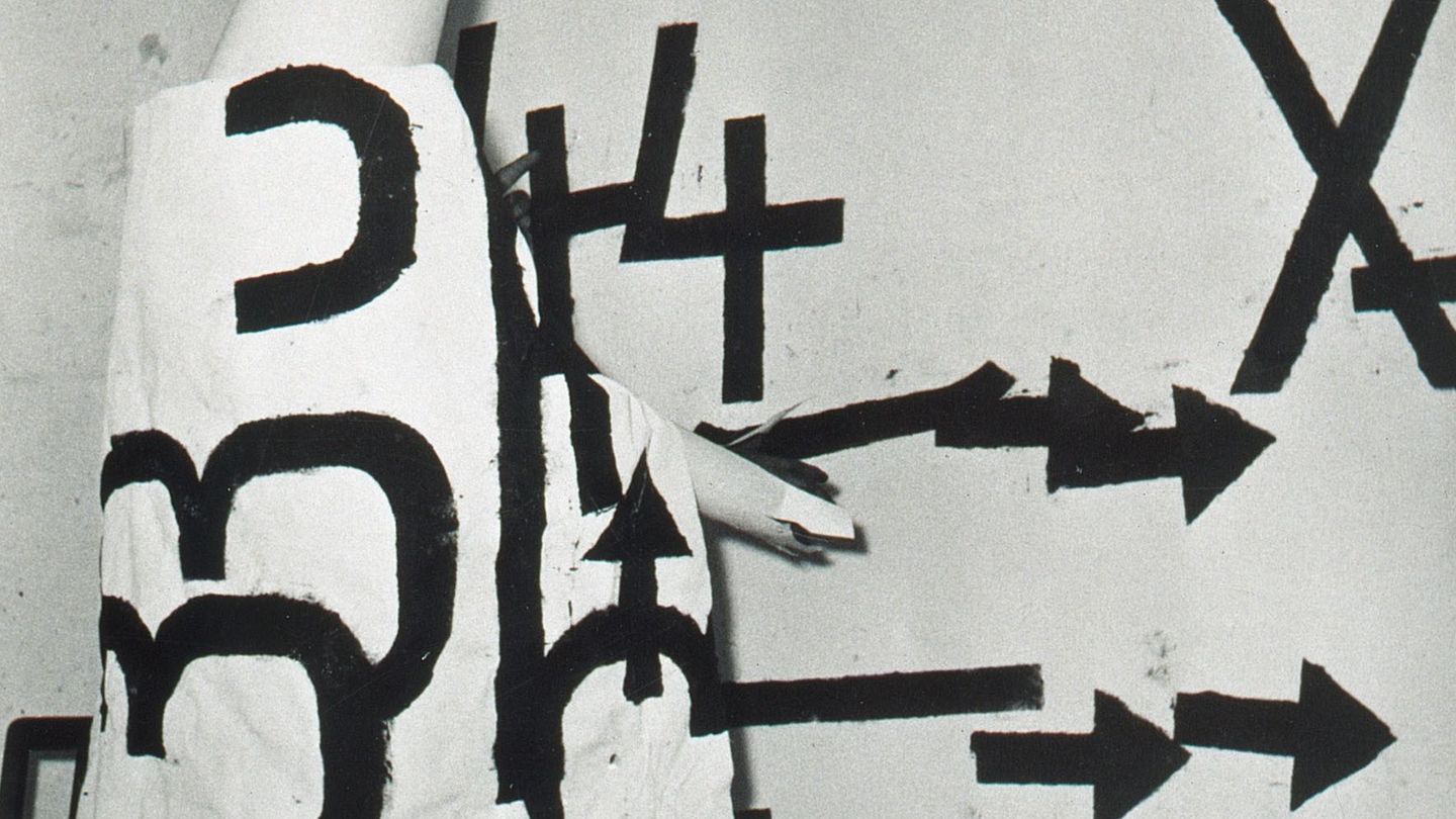 Photograph of Jannis Kounellis at a performance dressed in a white robe on which large black letters have been painted. Here he is in front of his own work Senza Titolo, which has the same painting style as the one on the robe. Claudio Abate, Sammlung Goetz Munich