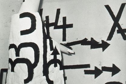 Photograph of Jannis Kounellis at a performance dressed in a white robe on which large black letters have been painted. Here he is in front of his own work Senza Titolo, which has the same painting style as the one on the robe. Claudio Abate, Sammlung Goetz Munich