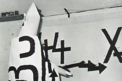 Fotografie von Jannis Kounellis bei einer Performance in eine weiße Kutte gekleidet, auf der schwarze große Buchstaben gemalt worden sind. Dabei befindet er sich vor seiner eigenen Arbeit Senza Titolo, die die gleiche Malweise wie die auf der Kutte aufweist. Claudio Abate, Sammlung Goetz München