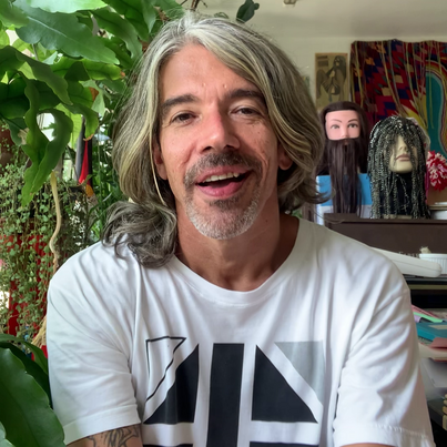 Der Künstler sitzt in einem Innenraum, von der Decke hängen Planzen im Hintergrund stehen Mannequin-Köpfe mit Perrücken in einem Regal