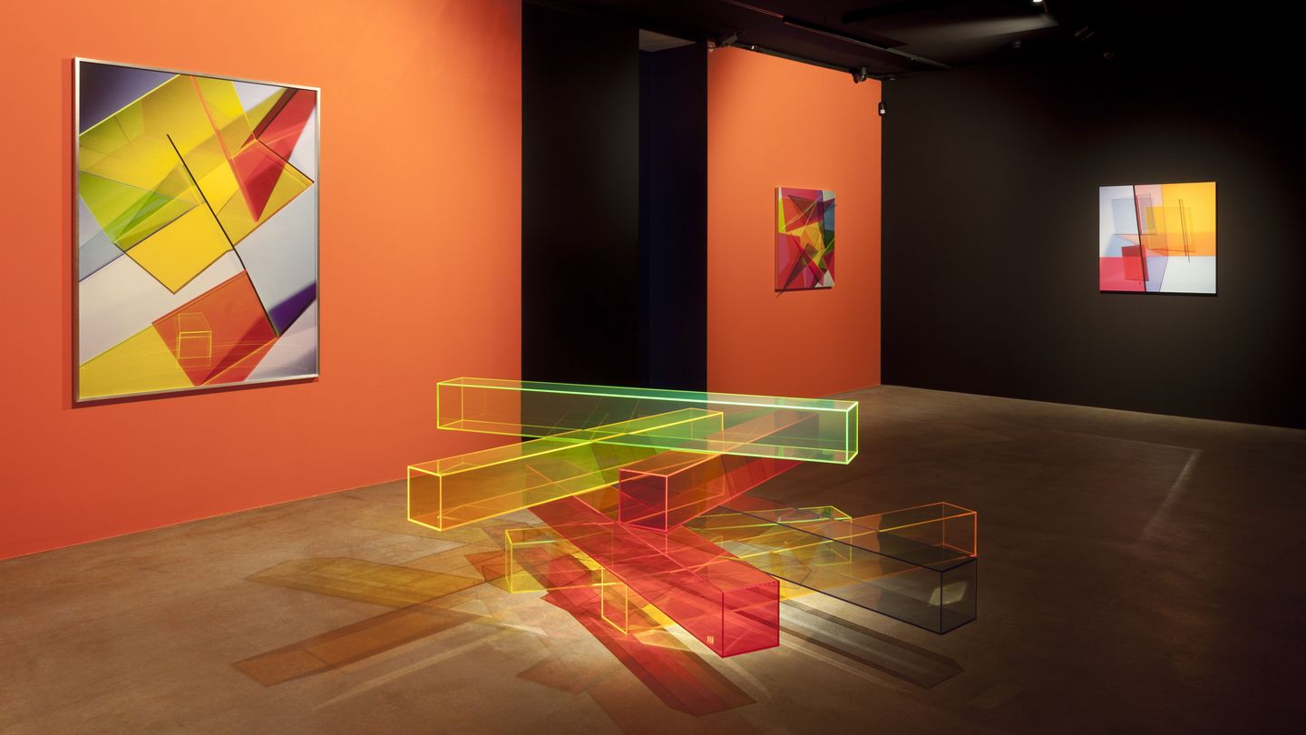 Exhibition view with geometric abstract photographs on orange wall and floor sculpture made of colorful plexiglass