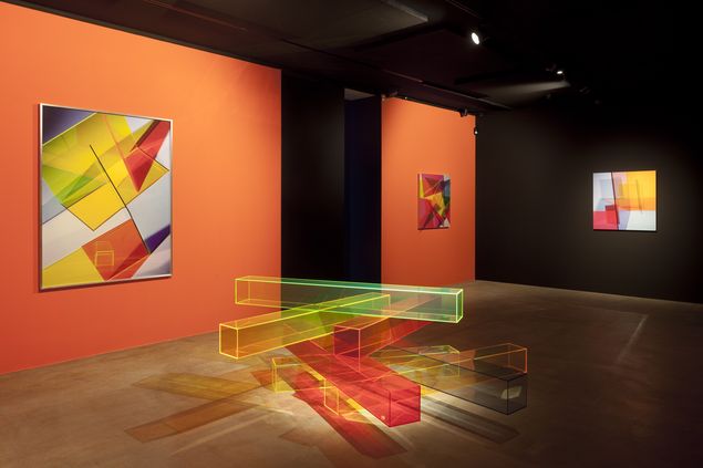 Ausstellungsansicht mit geometrisch abstrakten Fotografien an orangefarbiger Wand und einer Bodenskulptur aus buntem Plexiglas