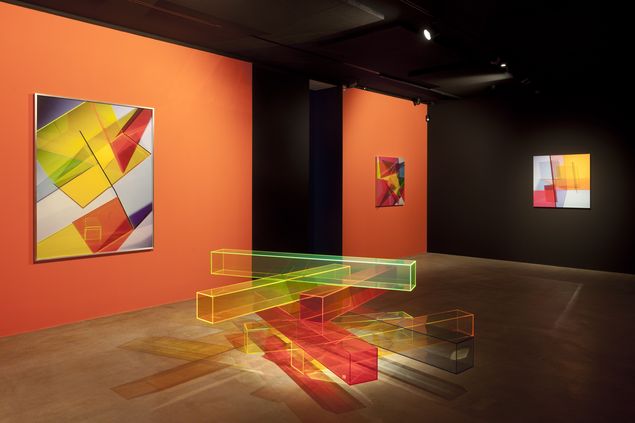 Exhibition view with geometric abstract photographs on orange wall and floor sculpture made of colorful plexiglass