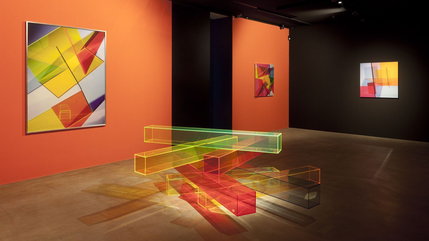 Ausstellungsansicht mit geometrisch abstrakten Fotografien an orangefarbiger Wand und einer Bodenskulptur aus buntem Plexiglas