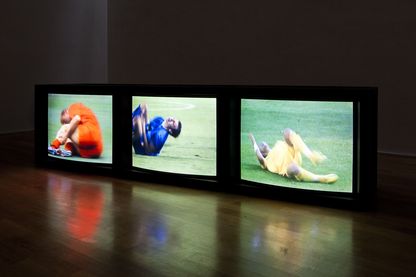 Videoinstallation von drei auf dem Boden stehenden, nebeneinander aufgereihten Monitoren, die alle drei verschiedene Szenen stürzender Fußballspieler in Nahaufnahme zeigen. Paul Pfeiffer, Sammlung Goetz München