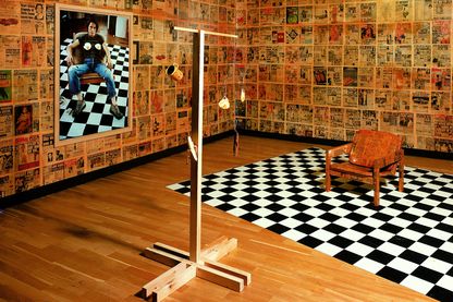 Hier sieht man einen ausgestalteten Raum der Künstlerin Sarah Lucas. Die Wände sind plakatiert mit Zeitungsseiten, an der linken Wand hängt ein fotografisches Selbstportrait mit Spiegeleiern auf der Brust platziert. In dem Selbstbildnis sitzt sie auf einem Stuhl, der auf einem schachbrettartigen Untergrund steht. Diese zwei Gegenstände finden sich auch in dem Raum wieder, der Stuhl ist nun auch mit Zeitungsausschnitten versehen worden. Davor befindet sich eine Art Mobile mit Spiegeleiern, Tasse, Banane und alter Bananenschale.