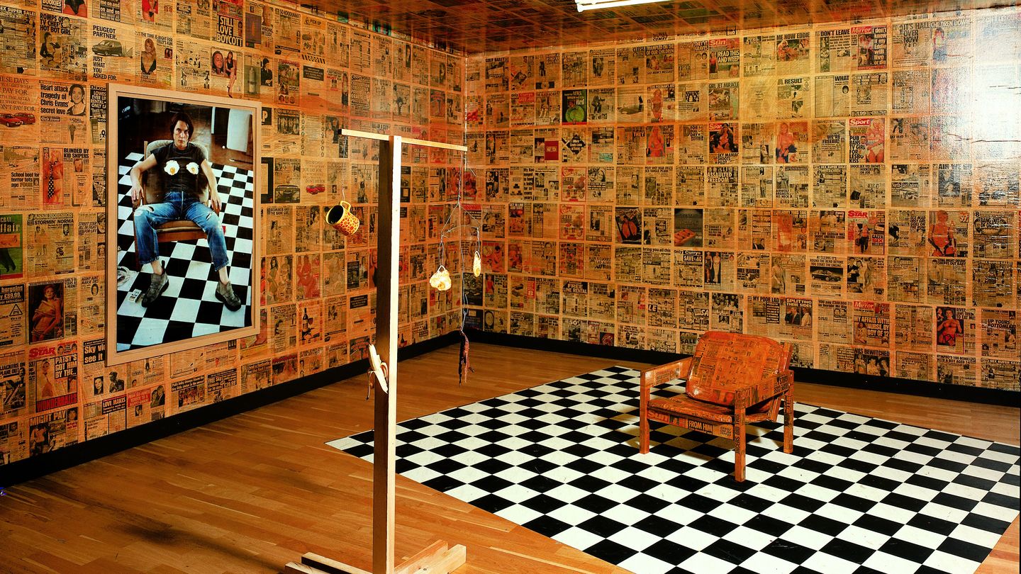 Here you can see a fully decorated room of the artist Sarah Lucas. The walls are covered with posters of newspapers, on the left wall hangs a photographic self-portrait with fried eggs placed on her chest. In the self-portrait she is sitting on a chair which is standing on a chessboard-like background. These two objects can also be found in the room, the chair has now also been provided with newspaper cuttings. In front of it there is a kind of mobile with fried eggs, cup, banana and old banana skin.