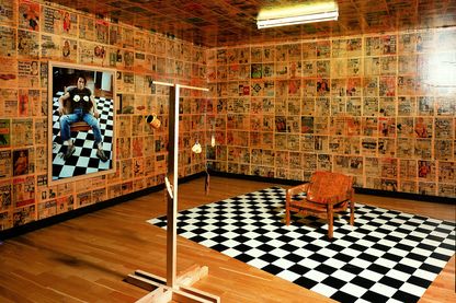 Here you can see a fully decorated room of the artist Sarah Lucas. The walls are covered with posters of newspapers, on the left wall hangs a photographic self-portrait with fried eggs placed on her chest. In the self-portrait she is sitting on a chair which is standing on a chessboard-like background. These two objects can also be found in the room, the chair has now also been provided with newspaper cuttings. In front of it there is a kind of mobile with fried eggs, cup, banana and old banana skin.
