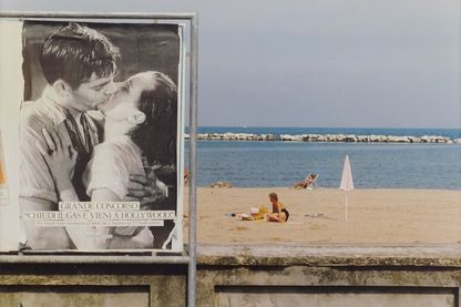Fotografie einer Strandszenerie mit einem Schwarzweiß-Plakat eines sich küssenden Paares davor. Luigi Ghirri, Sammlung Goetz München