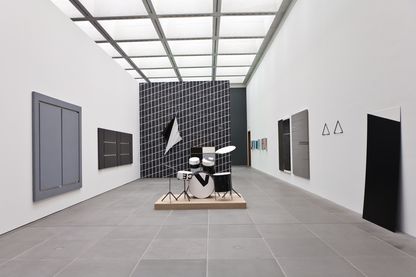 Ausstellungsansicht mit Arbeiten, die ausschließlich in den Farben Schwarz, Weiß und Grau gehalten sind. Dabei handelt es sich um bemalte oder bedruckte Leinwände, einer Tapete, einem Schlagzeug und Wandobjekten. Martin Boyce/Alan Charlton/Wade Guyton/Reiner Ruthenbeck/Michael Sailstorfer, Sammlung Goetz München