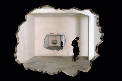 Videoprojektion, die ein Loch in einer Wand zeigt, hinter der eine Person in schwarz gekleidet steht. Hinter ihr befindet sich wieder ein Loch in einer Wand mit einer Person dahinter. Diese Situation scheint sich unendlich zu wiederholen. Zilla Leutenegger, Sammlung Goetz München