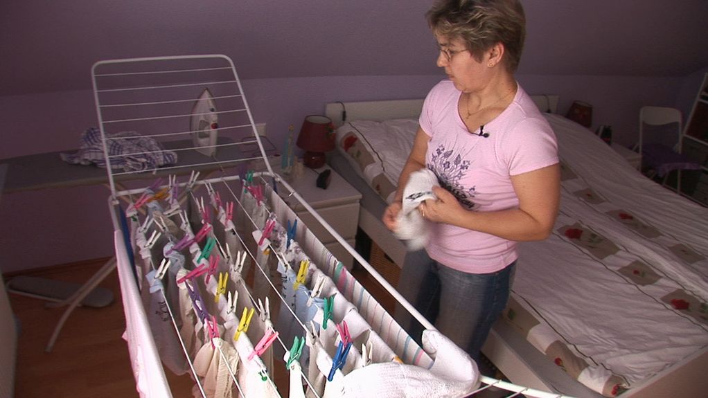 Das Video Still zeigt eine Frau mit kurzen, blonden Haaren in einem einfach eingerichteten Schlafzimmer vor einem bereits behängten Wäscheständer beim Wäsche aufhängen. Artur Żmijewski, Sammlung Goetz München