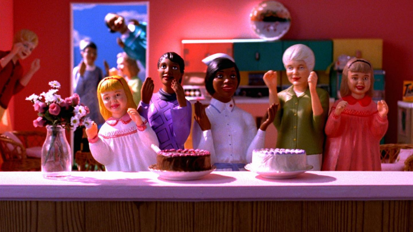 Playmobilfiguren im Puppenhaus hinter einem Tresen auf dem zwei Torten und eine Blumenvase stehen. Alle haben erhobene Hände und lächelnde Gesichter. Im Hitergund schauen weitere Figuren durch eine Tür