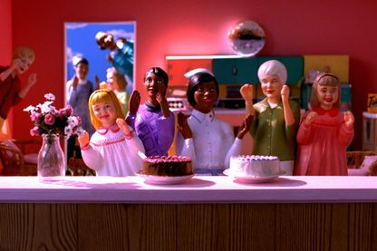 Playmobilfiguren im Puppenhaus hinter einem Tresen auf dem zwei Torten und eine Blumenvase stehen. Alle haben erhobene Hände und lächelnde Gesichter. Im Hitergund schauen weitere Figuren durch eine Tür
