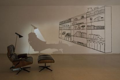 Installationsansicht, auf der ein schwarz gepolsterter Sessel mit einem gepolstertem Fußhocker zu sehen ist, eine Stehlampe spendet Licht im sonst abgedunkelten Raum. Rechts davor befindet sich ein gezeichnetes Regal, mit den darin befindlichen Büchern, Ordnern und Gefäßen. Hinter dem Sessel an der Wand ist ein Schatten desselbigen und der Stehleuchte auszumachen, außerdem scheint eine Person auf Sessel und Fußhocker zu liegen. Zilla Leutenegger, Sammlung Goetz München