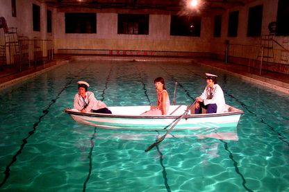 Auf dem Video Still sind drei asiatisch aussehende Personen in einem Boot sitzend zu sehen. Das Boot selbst befindet sich auf dem türkisblauen Wasser eines Schwimmbeckens in einem Schwimmbad. Yang Fudong, Sammlung Goetz München