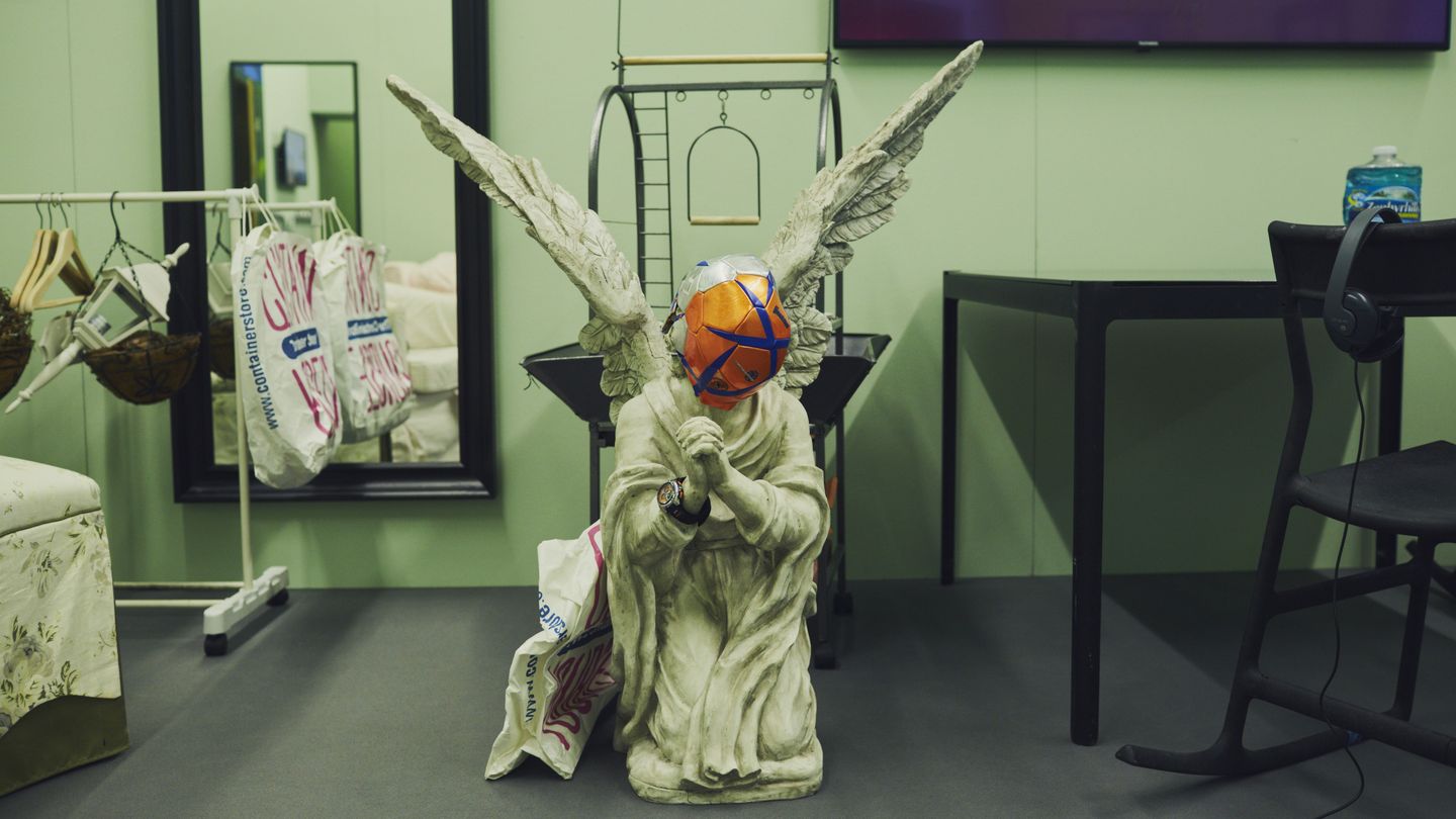 Detail of a stage set showing the statue of a praying funeral angel in the midst of living room furniture. On his face is one half of an orange football. Ryan Trecartin/Lizzie Fitch, Sammlung Goetz Munich