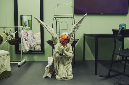 Detailansicht eines Bühnenbilds, das die Statue eines betenden Grabesengels inmitten von Wohnmöbeln zeigt. Auf seinem Gesicht befindet sich eine Hälfte eines orangenen Fußballs. Ryan Trecartin/Lizzie Fitch, Sammlung Goetz München