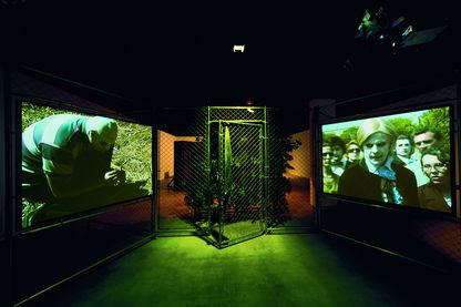 Installationsansicht der Arbeit "Woods Group - Extracurricular Activity Projective", bestehend aus einem Maschendrahtzaun mitsamt offenstehender Tür in der Mitte, daneben sind Monitore in den Maschendraht gehängt, die eine Videoarbeit Kelleys wiedergeben. Dahinter befindet sich ein Gestrüpp, in dem eine blonde Perücke auf einem Baumstumpf liegt. Der Raum ist abgedunkelt, eine Lichtquelle scheint auf die Tür im Maschendrahtzaun. Mike Kelley, Sammlung Goetz München