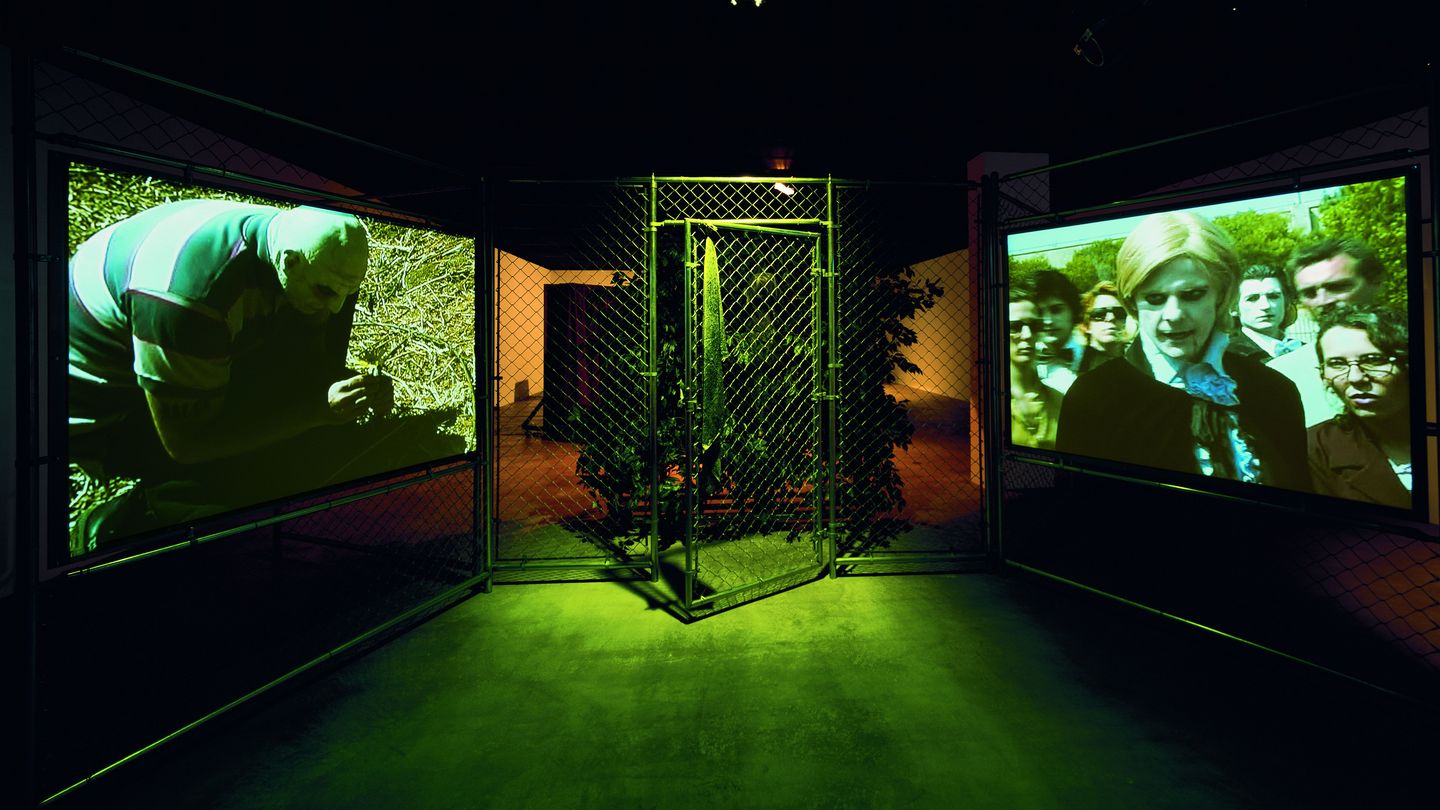 Installation view of the work "Woods Group - Extracurricular Activity Projective", consisting of a wire mesh fence including an open door in the middle, next to which monitors are hung in the wire mesh, reproducing a video work by Kelley. Behind it is a thicket in which a blond wig lies on a tree stump. The room is darkened, a light source shines on the door in the chain-link fence. Mike Kelley, Sammlung Goetz Munich