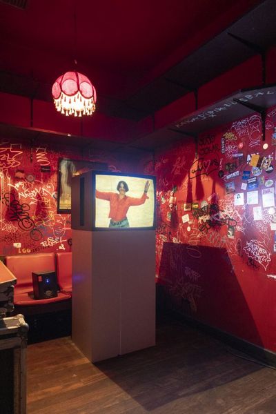 Darkened room with red walls covered with graffiti and inscriptions. In the center of the room is a pedestal with a monitor. On it is a woman with short dark hair, her arms raised in a dancing motion. Above the monitor hangs a lamp with an old-fashioned red fabric lampshade 