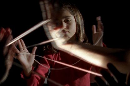 Dieses Filmstill zeigt ein junges Mädchen mit dunklen Haaren in einem dunklen Raum, das ein Spiel mit Fäden zwischen den Fingern spielt. Es scheint isoliert von den anderen mitspielenden Kindern zu sein, von denen man nur die erhobenen Hände mit den gleichen Fäden darin sieht. Doug Aitken, Sammlung Goetz München