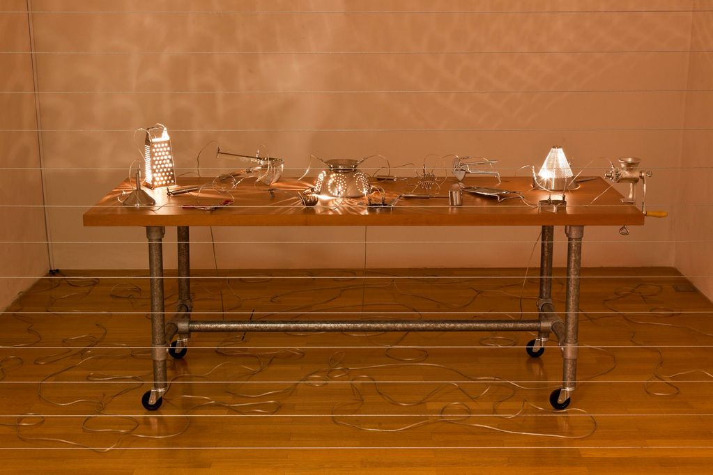Behind a wall of horizontally drawn transparent electric cables is a table on wheels with metallic kitchen utensils (e.g. pasta sieve, cheese grater, meat hammer), which are connected to each other by transparent electric cables and partly shine yellow. The floor is covered by the same cables. Mona Hatoum Collection Goetz, Munich 