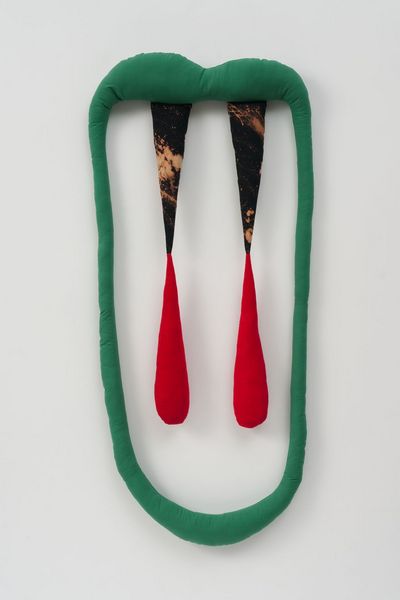 A green, padded fabric strip forms an oval, mouth-like shape. From the upper strip, which curves slightly like lips, additional filled fabric strips hang down, their tips tinted red and reminiscent of fangs.