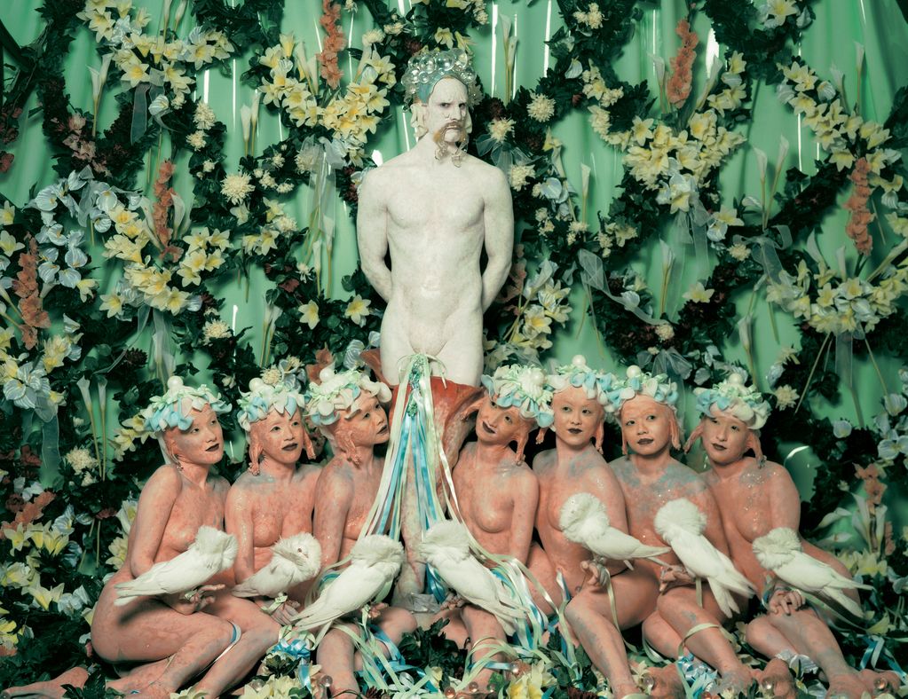 Hier sieht man ein Production Still aus der Cremaster-Reihe des Künstlers Matthew Barney. Vor einer Blumenwand steht ein Mann, weiß angemalt und einem Faun ähnlich sehend, vor ihm sitzen sieben unbekleidete Frauen mit seltsam anmutenden Ohren und Kopfschmuck. Sie halten jeweils eine weiße, seltene Taubenart auf ihrer Hand.