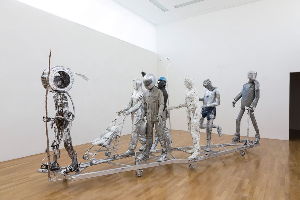 Life-size sculpture of a group of people made of metal, everyday objects (e.g. baby stroller, washing machine drum, clothes) on a mobile frame. Pawel Althamer, Sammlung Goetz, Munich 