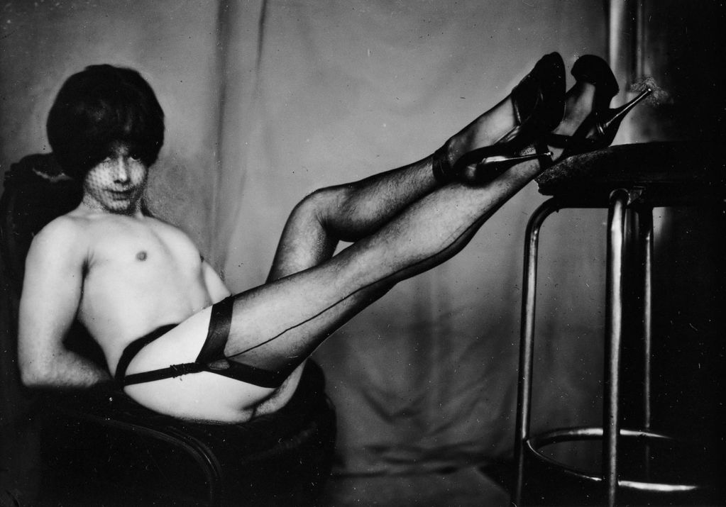 This black and white photography includes a complete shot of a man with face net and wig, garter belt and stockings as well as high heels, who looks lasciviously into the camera. He sits with his arms behind his back on a cantilever chair and has put his feet on a bar stool.